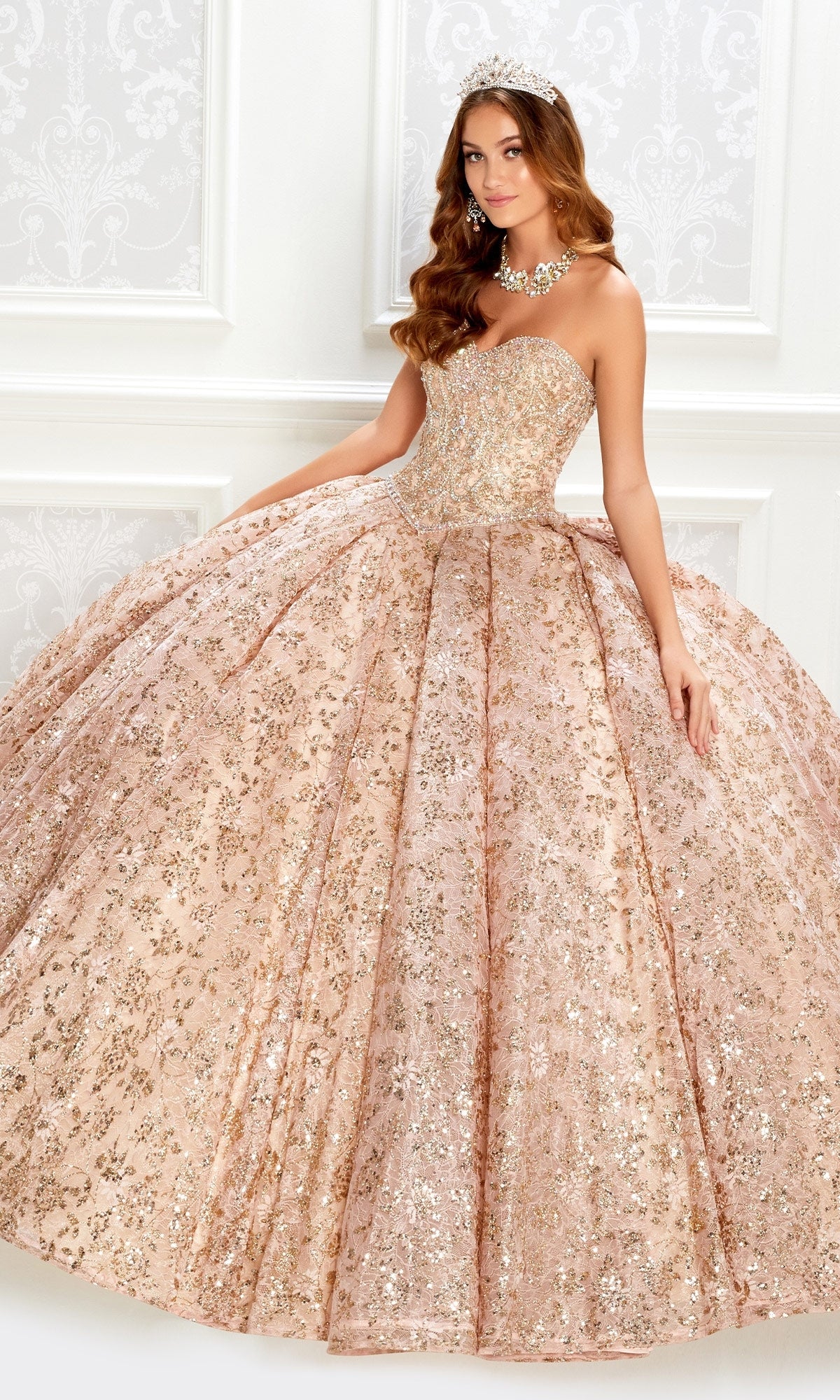A woman stands elegantly against a white ornate background, wearing the Strapless Quinceanera Dress: Princesa PR22022 with gold sequins and a full skirt, paired with a tiara and statement necklace.
